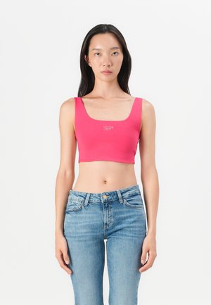 Tettsittende rosa crop-top med brede stropper og rund halsringning. Lett, elastisk stoff med en liten sølvlogo på forsiden. Kombinert med blå jeans.