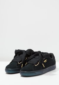 Baskets Puma en daim noir avec des lacets noirs épais, des œillets dorés, un logo doré et une semelle noire texturée, placées côte à côte sur une surface blanche.