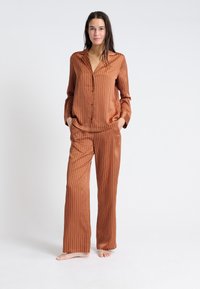 Zestaw piżamowy w kolorze rdzy z pionowymi paskami. Koszula ma kołnierzyk i zapięcie na guziki, natomiast spodnie są szerokie z elastycznym pasem.