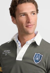 Polo color verde oliva con acentos en blanco en el collar y los puños, con un logo bordado en el pecho y una tapeta con botones.