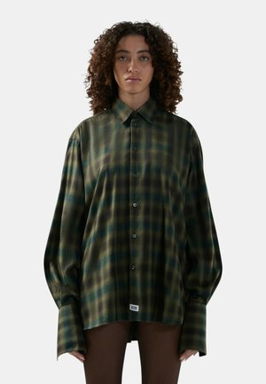 Persona con capelli ricci che indossa una camicia a quadri verde e nera oversize con maniche prolungate e pantaloni marroni su sfondo semplice.