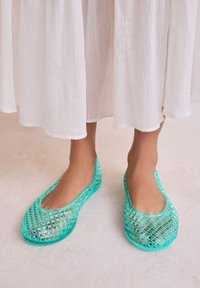 Poros turkio spalvos tinklelio, be raištelių batų su apvalia nosimi ir iškirptu dizainu. Dėvimi su balta, laisvai krentančia suknele, padėti ant šviesios paviršiaus.