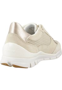 Zapatilla de deporte de ante y malla color beige con detalles metálicos, suela blanca, logotipo trasero y detalles texturizados. Presenta una forma redondeada y un collar acolchado.
