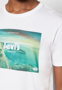 Camiseta de algodón blanca con un gráfico colorido que representa una escena costera, con el texto "Levi's" y "World Wide" en negrita. Mangas cortas, cuello redondo.
