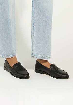 Black leather loafers featuring a flat sole, stitched detailing, and a wide strap across the instep, paired with light-washed denim.
