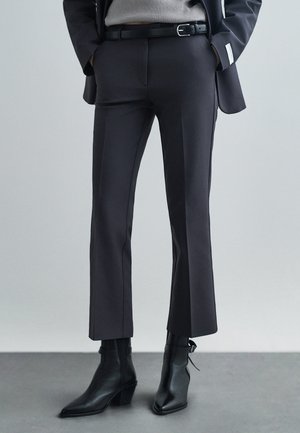 Calças pretas ajustadas com corte reto, apresentando uma textura elegante e detalhes mínimos, acompanhadas de botas de tornozelo pretas e pontiagudas.