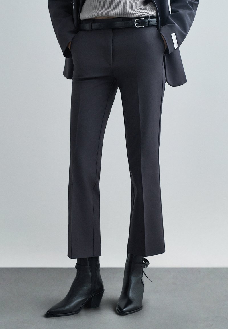 Mustad kohandatud püksid sirge lõikega, millel on sujuv tekstuur ja minimaalne detailide arv, koos terava ninaga mustade pahkluu saabastega.