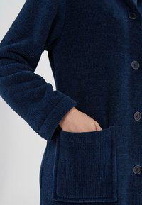 Chaqueta de canalé azul marino con cuello puntiagudo, botones frontales y un bolsillo lateral. La textura es suave y de punto, con un corte holgado.