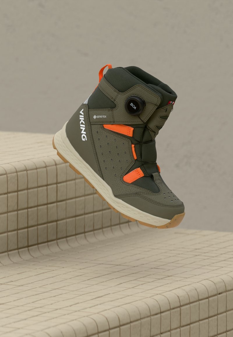 Aukštos olivinės žalios ir oranžinės Viking Gore-Tex batai su BOA dalykų raišteliu, rodoma pakabinta virš smėlio spalvos plytelių laiptų.