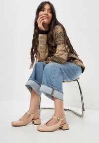 Beige patent slingback -kengät, joissa on T-nauha, neliönmuotoinen kärki ja matala lohko-osa, yhdistettynä sinisiin leveälahkeisiin farkkuihin ja teksturoituun neuletakkiin.