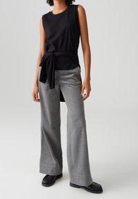 Top noir sans manches avec un nœud à la taille, associé à un pantalon en velours côtelé gris à jambes larges et des chaussures noires, avec une finition texturée.