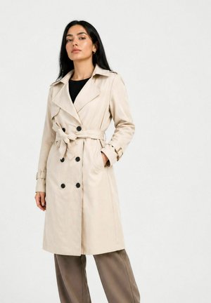 Kvinde stående iført en beige trenchcoat med bælte over en sort top og brune vidde bukser mod en ensfarvet lys baggrund.