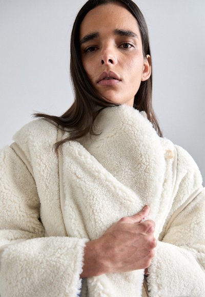 Palton din fleece, de culoare crem, pufos, cu un design tip wrap; dispune de un buton vizibil și mangas largi; textură moale.