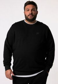 Sudadera negra de cuello redondo con un acabado texturizado, que presenta un pequeño logo oscuro en el pecho, superpuesta sobre una camisa blanca.