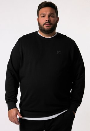 Zwarte crewneck sweatshirt met een gestructureerde afwerking, voorzien van een klein, donker logo op de borst, gelaagd over een wit shirt.