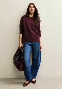 Maglione borgogna, jeans a gamba larga blu, borsa lucida scura, ballerine color maroon, su uno sfondo neutro. Collana semplice e capelli lisci.