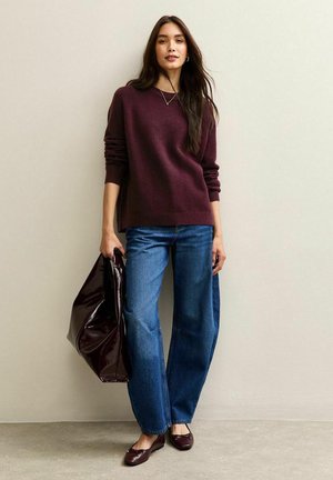 Maglione borgogna, jeans a gamba larga blu, borsa lucida scura, ballerine color maroon, su uno sfondo neutro. Collana semplice e capelli lisci.