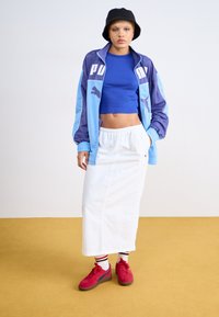 PUMA vindjacka i blått med marinblå detaljer över en blå crop top, tillsammans med en vit kjol och röda sneakers. Svart bucket hat.