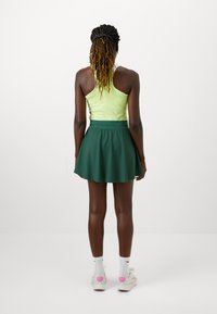 Groene skaterrok met een aansluitende tailleband, gecombineerd met een felgele tanktop. Het model draagt witte sokken en een lichtgekleurde sneakers.