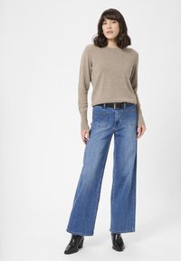 Kvinde står iført en beige langærmet sweater, blå højtaljede bukser med brede ben og forreste lommer, sort bælte og sorte ankelstøvler.