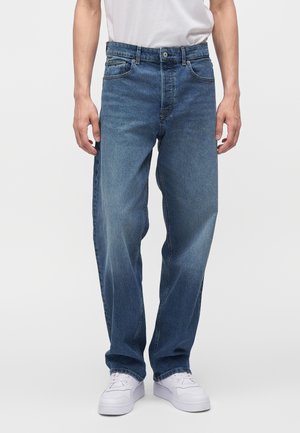 Mand iført medium blå jeans med lige ben, hvide sneakers og en hvid t-shirt, stående mod en ensfarvet baggrund.