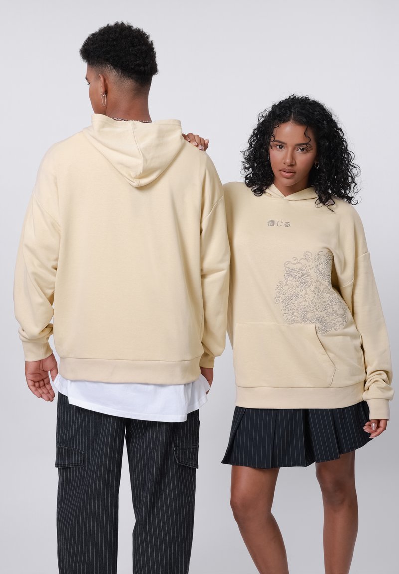 Prehnaný béžový sweater s predným vreckom, so kvetinovým motívom na boku. Vybavený čiernymi prúžkovanými nohavicami a plisovanou sukňou.