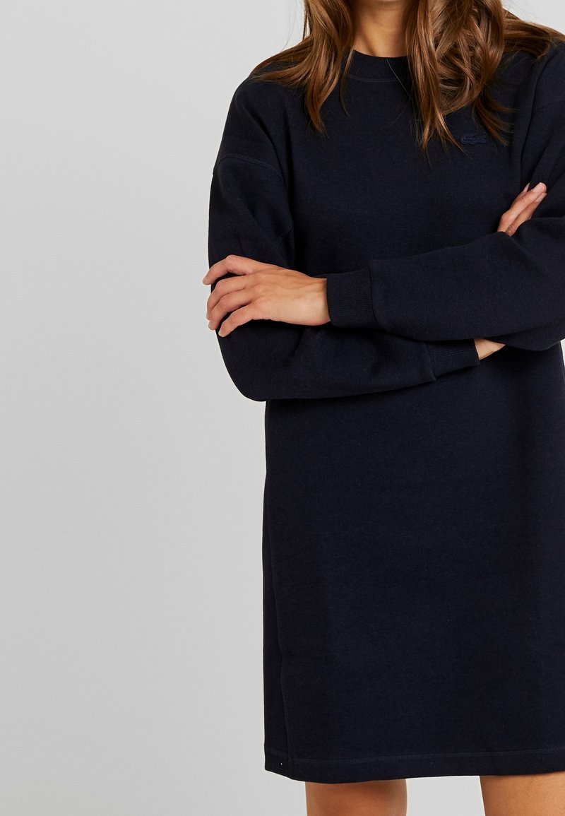 Femme portant une robe pull à manches longues bleu marine foncé, bras croisés devant un fond gris clair uni.