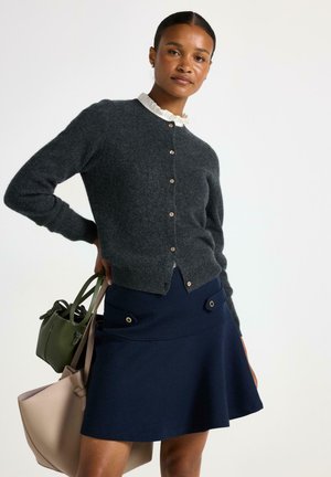 Donna che indossa un cardigan grigio scuro con bottoni, una blusa bianca con volant, una gonna blu navy, mentre tiene delle borse beige e verde su uno sfondo neutro.