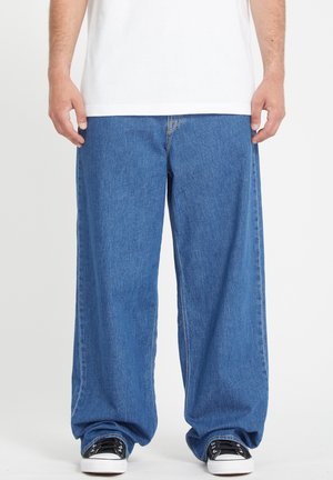 Blaue weit geschnittene Jeans aus Denim, mit lockerer Passform, kontrastierenden Nähten und klassischem Fünf-Taschen-Design. Getragen mit schwarzen Sneakers.