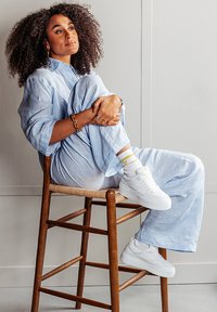 Light blue linen two-piece outfit, featuring wide-leg pants and a long shirt. White sneakers with yellow accents, sitting on a wooden stool.