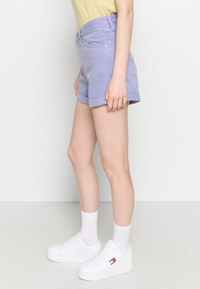 Persoon in lichtpaarse korte broek met omgeslagen zoom, witte sokken en witte sneakers met een rood, wit en blauw logo, staand tegen een effen achtergrond.