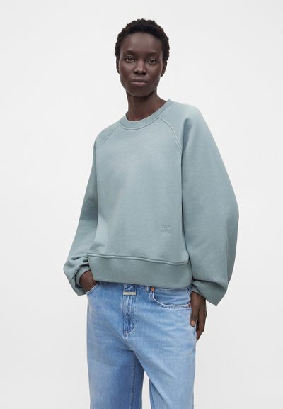 Femme aux cheveux courts portant un sweat-shirt bleu clair ample et un jean bleu clair, debout les mains dans les poches devant un fond blanc.