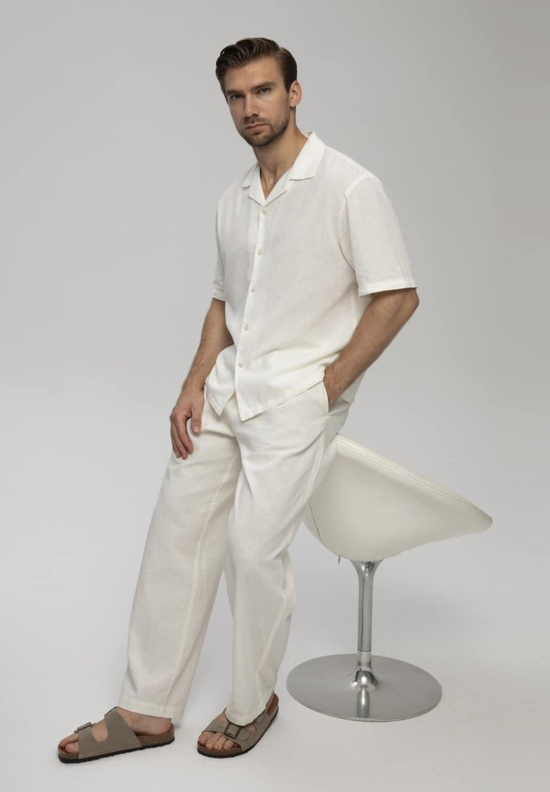 Uomo con camicia bianca a maniche corte e pantaloni seduto su una moderna sedia girevole bianca, indossa sandali marroni su sfondo uniforme.