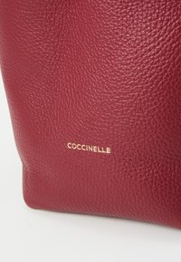 Rød læder håndtaske med struktureret overflade, der har et guld "Coccinelle" logo. Kompakt design med bløde kanter og ingen synlig hardware.