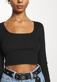 Nainen, joka on pukeutunut mustaan pitkähiharaiseen cropped-toppiin, jossa on neliökaula-aukko, sekä vaaleansininisiin farkkuihin, joissa on musta vyö, jossa on hopeiset reiät.