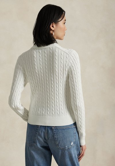 Hvit kabelstrikket genser med ribbestrikket kant og mansjetter, med et teksturert mønster. Brukt med blå slitte jeans, sett bakfra.