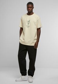 Beige bomull t-skjorte med grafisk trykk av en engel og teksten "Lucid Lights" i grønt, sammen med svarte joggebukser og hvite sneakers.