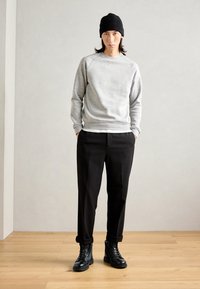 Vyras, dėvintis juodą kepurę, pilką sportinį megztinį, juodas kelnes su lankeliais ir juodus odinius batus, stovi ant medinių grindų priešais vientisą sieną.