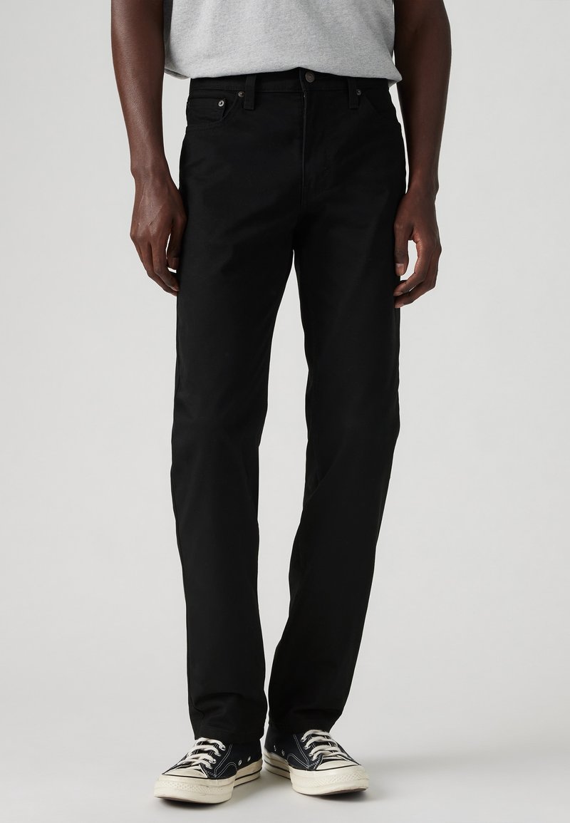 Pantalones de algodón negros con corte recto, diseño de cinco bolsillos y detalles sutiles de costura; combinados con zapatillas negras y blancas.