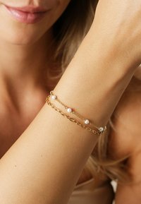 Guldkedja med små vita pärlor, med eleganta kedjor och en växelvis länkdesign. Slät textur på en ljus hudton.