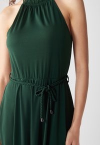 Kvinna i en mörkgrön ärmlös halterneckklänning med ett flätat bälte knytet i en rosett vid midjan, visad från halsen till höfterna.