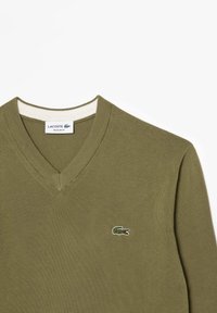 Suéter en verde oliva con cuello en V, confeccionado en material de punto texturizado, que presenta un acento blanco en el cuello y un pequeño logo bordado de un cocodrilo.