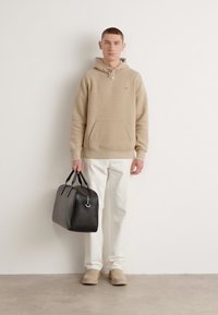 Beige hoodie with a front pocket, white pants, and tan slippers. Holding a black leather duffel bag with a silver hardware detail.