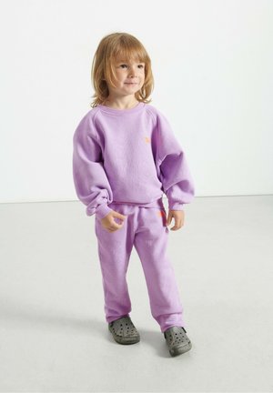 Lilla puuvillane sviiter ja sobivad püksid elastsete mansettidega; rinnal on oranž logo, kantud mustriliste hallide puukingadega.