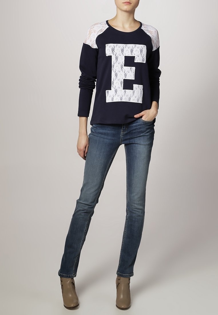 Maglione navy con pannelli in pizzo bianco, grande grafica "E", e maniche lunghe, abbinato a jeans azzurri chiari e stivaletti beige.