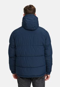 Mężczyzna w granatowej kurtce pikowanej z kapturem, widok z tyłu, stojący na tle jasnym.