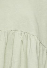 Světle zelený top s měkkou texturovanou tkaninou a nařaseným volánem na spodním lemu, který vytváří vrstvený design.