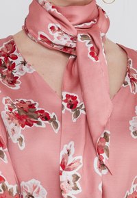 Sciarpa in satin rosa con motivo floreale, fiori rossi e bianchi, annodata intorno al collo, e una blusa a V con un design floreale simile.