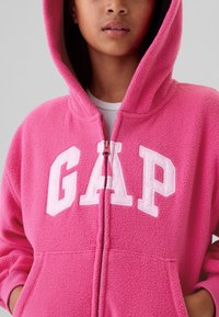 Mladostna oseba, oblečena v svetlo roza flis s kapuco z zadrgo spredaj in velikimi belimi črkami "GAP" na prsih.