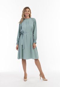 Rochie de culoare teal deschis, cu mâneci lungi, cu manșete plisate și un cordon asortat. Material neted, lungime până la genunchi și decolteu rotund. Croi clasic.
